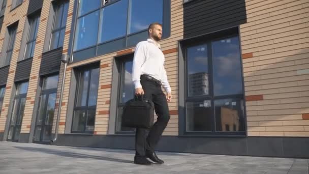 現代的なオフィスビルの近くを歩くブリーフケース付きの成功したビジネスマン。若い自信に満ちた起業家が歩道で仕事をする予定です。仕事に通勤するハンサムな男性マネージャー。スローモーション — ストック動画