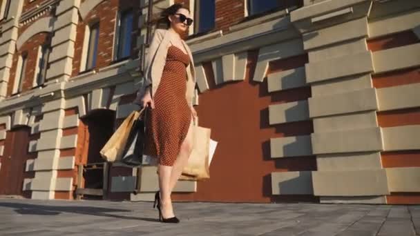 Attractive young lady holds shopping bags walking at sidewalk after purchases. Fashionable woman carries paper packages going along urban street. Concept of leisure and free time. Low view Slow motion — Stock Video