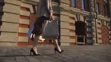 Şehir caddesinde yürüyen genç kadınların kolları alışveriş çantaları taşıyor. Paketlerle şehir meydanına giden bir bayan. Siyah ayakkabılı, yüksek topuklu bir kadın kaldırıma çıkıyor. Dolly vurdu.