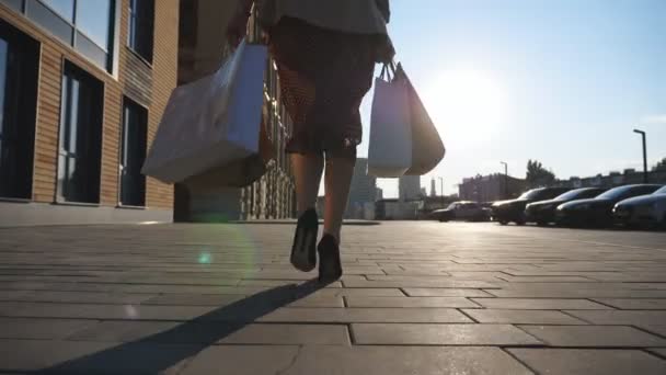 Pies femeninos en zapatos negros en tacones altos pisando la acera. Chica va con paquetes en la plaza de la ciudad después de las compras. Manos de mujer joven llevan bolsas de compras caminando por la calle urbana. Vista trasera — Vídeo de stock