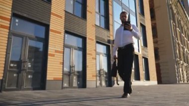 Resmi giyimde başarılı bir girişimci ofis binası yakınlarında akıllı telefon kullanır. Çantalı, cep telefonlu, kaldırımda işe giden kendinden emin genç bir işadamı. Dolly çekim ağır çekim