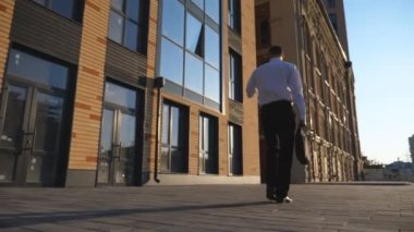 Evrak çantalı genç işadamı cep telefonuyla kaldırımda işe gidiyor. Başarılı bir girişimci resmi giyimde şehir caddesindeki ofis binasının yakınında akıllı telefon kullanıyor. Ağır çekim.