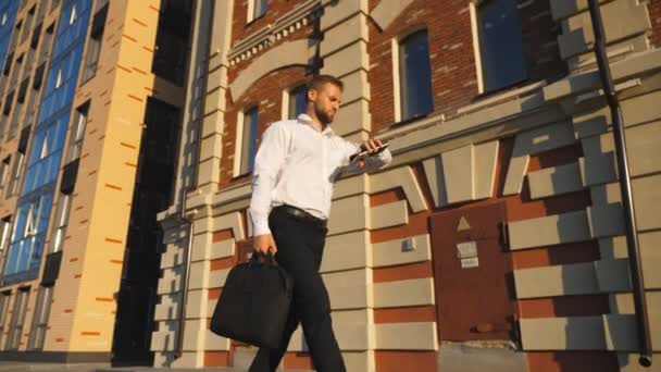 ブリーフケース付きの若いマネージャーは歩道のオフィスビルの近くを歩いている彼の時計を見ます。正式な着用で成功したビジネスマンは、街の通りで働くために行くスマートフォンを使用します。スローモーション — ストック動画