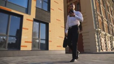Çantalı, cep telefonlu, kaldırımda işe giden kendinden emin genç bir işadamı. Resmi giyimde başarılı bir girişimci ofis binası yakınlarında akıllı telefon kullanır. Dolly çekim ağır çekim