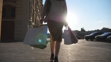 Şık bir kadının arka görünüşü, şehir caddesinde yürüyen kağıt paketler taşıyor. Zarif genç kız alışveriş sonrası kaldırıma çıkan alışveriş torbalarını tutuyor. Mevsimlik satış ve indirim kavramı. Yavaş çekim