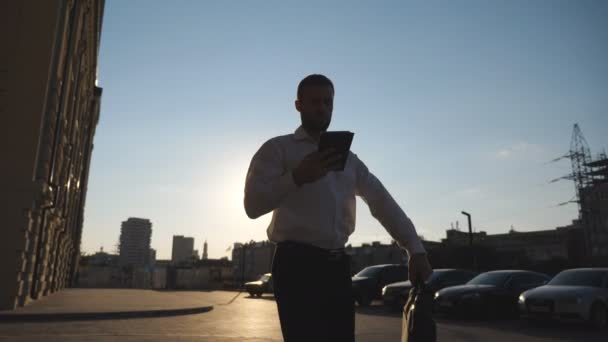Успешный бизнесмен в формальной одежде использует смартфон, идущий на работу на городской улице. Уверенный молодой предприниматель с портфелем смотрит на часы, идущие рядом с офисным зданием по тротуару. Долли выстрелил — стоковое видео