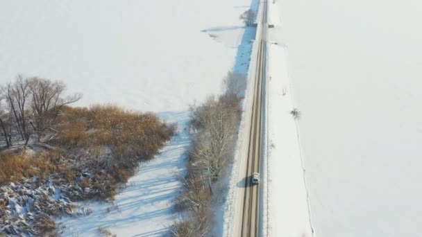 Plano aéreo de coche a caballo a través de carretera cubierta de nieve cerca del lago congelado. SUV blanco conduciendo en ruta de presa en el día de invierno. Volando sobre el auto moviéndose a través del puente del río. Camino paisajístico escénico. Vista superior — Vídeos de Stock