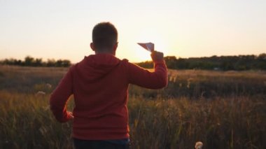 Çayırda kağıt uçakla koşan sevimli küçük bir çocuğun arka manzarası. Kaygısız küçük çocuk çim tarlasında koşuyor ve gün batımında oyuncak uçak fırlatıyor. Çocukluk hayali kavramı