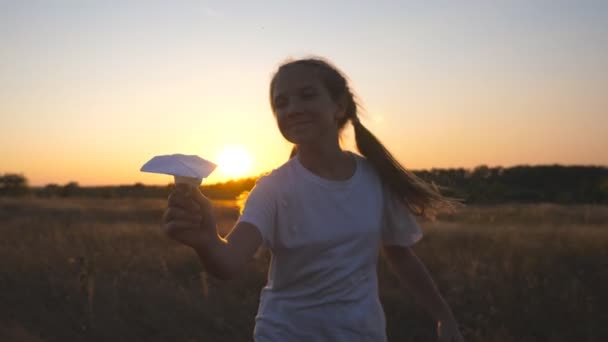 Szczęśliwa mała dziewczynka biegająca z papierowym samolotem przez trawiaste pole. Wesoły mały dziecko biegający z zabawkowy samolot wśród łąka nad zachodem słońca tło. Słodki dzieciak bawiący się w naturę. Koncepcja dziecięcych marzeń — Wideo stockowe