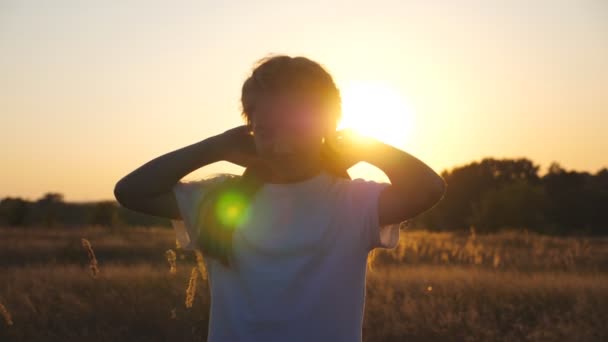 Glückliches kleines Kind blickt in die Kamera, steht auf der Wiese und spielt mit ihren Haaren. Porträt eines kleinen lächelnden Mädchens im Gras vor dem Hintergrund des Sonnenuntergangs. Konzept der Sorglosigkeit und Freiheit. Dolly erschossen — Stockvideo