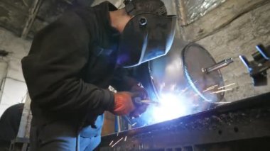 Metal parçalarını kaynaştıran işçiler. Kaynakçı iki çelik detay arasında ortaklık yapıyor. Metal kaynak yapan koruyucu maskeli bir adam. Endüstriyel üretim. Düşük açılı çekim