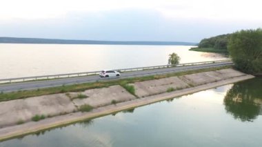 Göl kenarındaki yolda uçan arabanın görüntüsü. Beyaz SUV yaz günü baraj yolunda gidiyor. Otomatik olarak nehir ya da gölet köprüsünden geçiyor. Manzaralı bir yer. Seyahat ve tatil kavramı. Üst görünüm