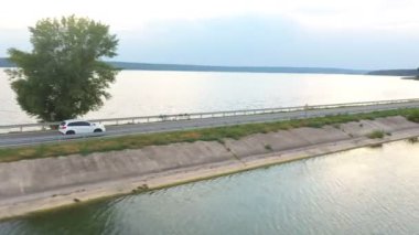 Göl kenarındaki yolda uçan arabanın görüntüsü. Beyaz SUV yaz günü baraj yolunda gidiyor. Otomatik olarak nehir ya da gölet köprüsünden geçiyor. Manzaralı bir yer. Seyahat ve tatil kavramı. Üst görünüm