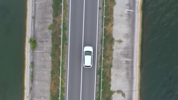 Blick von oben auf weißes Auto fährt durch Staudamm-Route. SUV fährt an einem Sommertag auf einer Straße in der Nähe des Sees. Der Flug über das Auto bewegt sich durch eine Brücke aus Fluss oder Teich. Landschaftlich reizvoller Weg. Reisekonzept. Luftaufnahme — Stockvideo