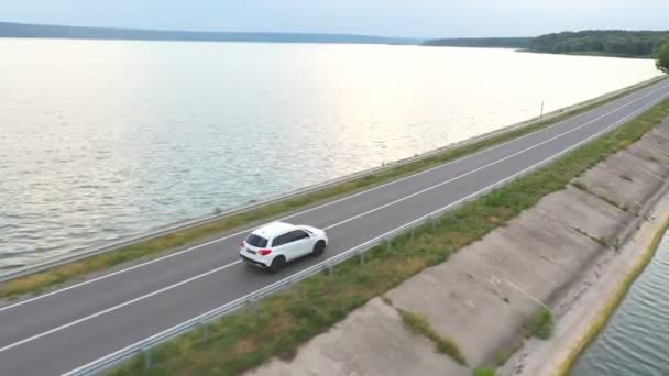 Aerial shot of white car rides through dam route on summer day. SUV drives at road near lake or pond. Flying over the auto moves through bridge of river. Concept of weekend journey or travel. Top view — ストック動画