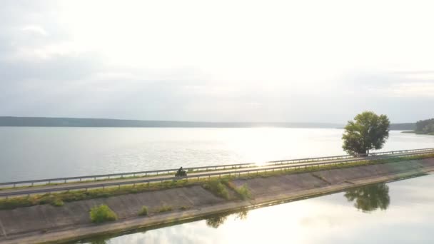 Légi kilátás motoros lovaglás a gát útvonalon napsütés a háttérben. Motoros versenyez a motorkerékpárjával az úton közel a tóhoz. Ember vezet biciklit utazás közben. A kaland fogalma. Felülnézet — Stock videók