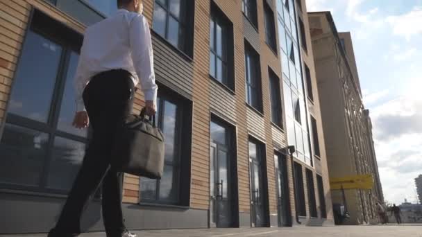 Mladý sebevědomý podnikatel ve formálním oblečení kráčí v blízkosti moderní kancelářské budovy. Úspěšný obchodník s aktovkou na chodníku. Manažer dojíždí do práce. Dolly shot Zpomalený pohyb — Stock video
