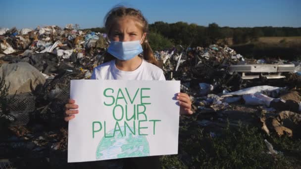 Porträt eines kleinen Mädchens in medizinischer Schutzmaske blickt vor dem Hintergrund einer Mülldeponie in die Kamera. Wenig Öko-Freiwillige halten Plakat der Umweltbewegung zur Rettung der Erde in den Händen. Dolly erschossen — Stockvideo