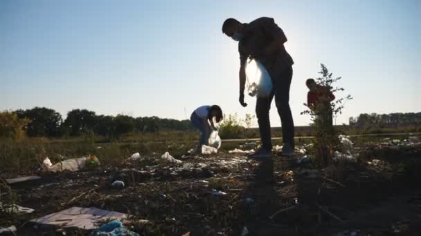 Family of eco activists in masks cleaning meadow of plastic and paper waste. Young parents with little son collecting trash in bags at lawn near roadside. Concept of environmental problem. Dolly shot — Stock Video