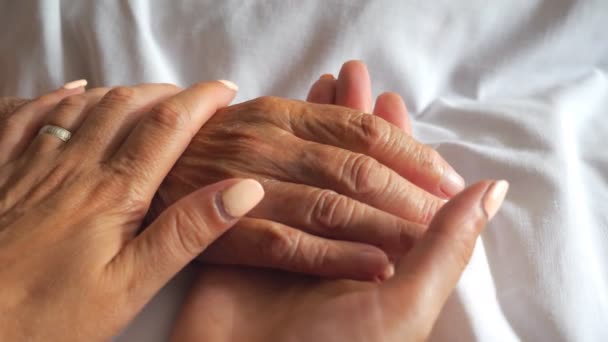 Young woman gentle stroking hand of mother taking care her. Daughter comforting wrinkled arm of elderly mom lying at bed in hospital. Girl showing love to parent. Detail view Slow motion — ストック動画