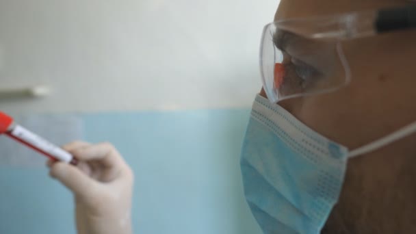 Profil des wissenschaftlichen Reagenzglases mit Blutprobe auf Coronavirus. Junge Laborarbeiterin in Schutzbrille bei der Untersuchung von Blutproben auf COVID-19. Sicherheit vor der Pandemie des Coronavirus — Stockvideo