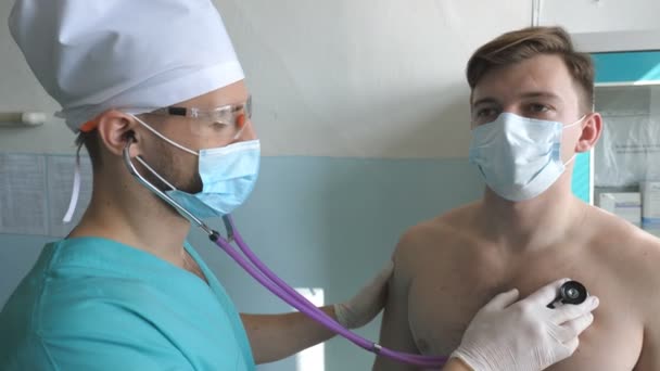 Medic met stethoscoop luistert longen van patiënt kijkt voor COVID-19 symptomen. Dokter met een onderzoekende man met stethoscoop aan coronavirus longontsteking tekenen. Artsen controleren van de borst van een man in het ziekenhuis — Stockvideo