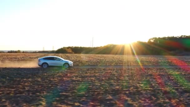 Tiro aéreo de um carro eléctrico a mover-se pela estrada com rasto de poeira atrás. Nova auto condução ao longo da estrada rural. Veículo moderno montando em carga elétrica. Paisagem cênica no fundo — Vídeo de Stock