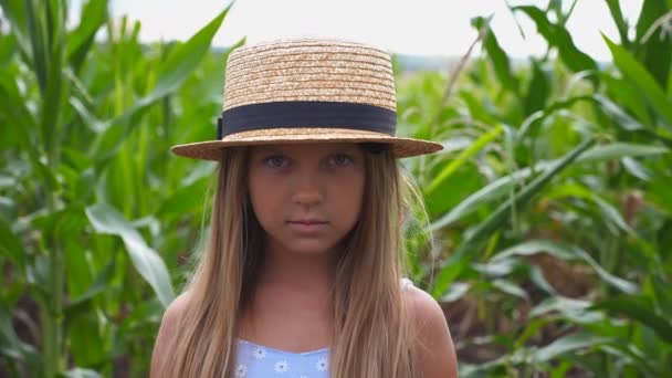 Portrait de petite fille sérieuse en chapeau de paille regardant dans la caméra sur le fond flou du champ de maïs à la ferme biologique. Petit garçon aux longs cheveux blonds debout dans la prairie. Gros plan — Video