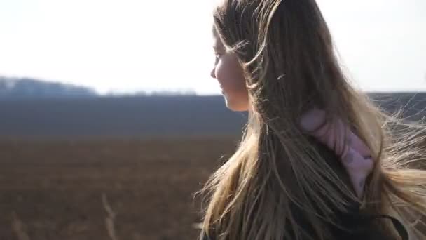 Klein lachend meisje met blaas blond haar wandelingen op de weg in de buurt van geploegd veld. Gelukkig kind met rugzak verhuist op weg naar school. Vrouwelijke kind gaat langs een oprit in de buurt van geploegde weide. Dolly schot — Stockvideo