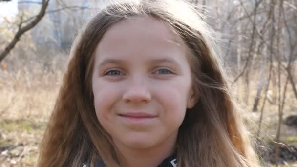 Retrato de un niño pequeño y feliz sobre el fondo borroso de un bosque a principios de primavera. Sonriente chica rubia mirando a la cámara al aire libre. Las emociones cercanas del chiquitín femenino con la expresión alegre en la persona — Vídeo de stock