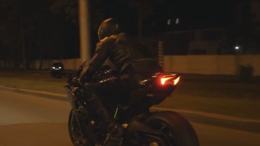 Motosikletli, akşam saatlerinde motosikletiyle yarışıyor. Şehrin gece caddesinde modern motosiklet süren bir adam. Yolculuk sırasında bisiklet süren adam. Özgürlük ve macera kavramı. Arka görünüm Kapat