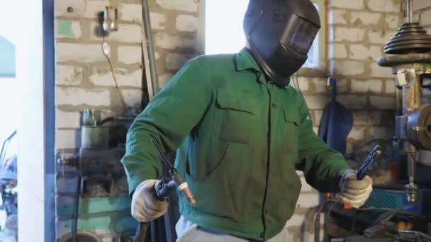 Cheerful male welder in protective mask and uniform dancing funny like a robot in garage. Happy mechanic with welding machine and pliers performing robotic moves to rhythm of music. Dolly shot — Stock Video