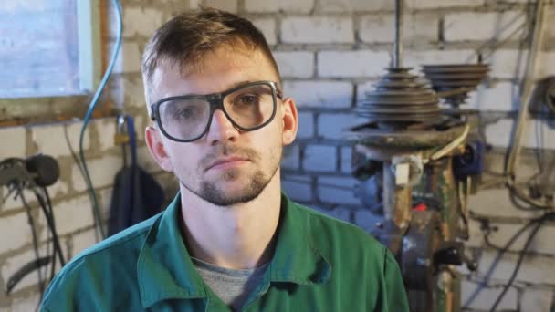 Vyčerpaný mladý opravář v ochranných brýlích, hledící do kamery v garáži. Portrét unaveného profesionálního mechanika v dílně. Koncept údržby. Dolly shot Close up — Stock video