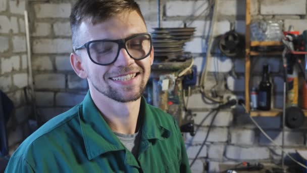 Glad ung mekaniker tar av sig skyddsglasögon efter att ha avslutat sitt jobb. Porträtt av reparatör i arbetskläder skrattar och tittar in i kameran. Leende ung man med skägg i garaget. Närbild — Stockvideo