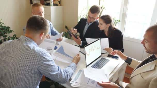 近代的なオフィスでのチームミーティング中に収入チャートやグラフを議論ビジネスの人々.若い同僚がテーブルに座って財務報告を分析しています。机の上で書類を調べる同僚 — ストック動画