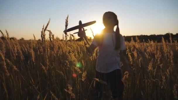 Szczęśliwa dziewczynka biegnąca samolotem przez trawiaste pole. Beztroskie dziecko biegnie wśród łąki bawiąc się samolotem zabawki na tle zachodu słońca. Małe dziecko bawiące się z naturą. Koncepcja dziecięcego snu — Wideo stockowe