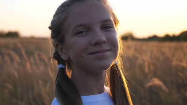Portret szczęśliwej uśmiechniętej dziewczyny patrzącej w kamerę na tle trawy. Małe dziecko stoi na letniej łące. Zbliż emocje małego dziecka z dobrą miną. Dolly strzał — Wideo stockowe