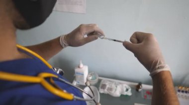 Tıbbi eldivenli ve maskeli doktor Covid-19 aşı sıvısını şırıngaya çekiyor. Erkek hastane çalışanı iğneye ilaç hazırlıyor. Laboratuvarda çalışan bir bilim adamı. Corona virüsü salgını kavramı.