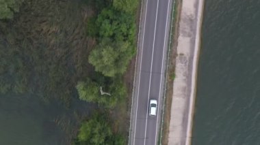 Baraj yolundan geçen beyaz arabanın üst görüntüsü. SUV yaz günü göl kenarındaki yolda gidiyor. Oto üzerinden uçmak nehir ya da gölet köprüsünden geçer. Manzaralı bir yer. Seyahat konsepti. Hava görüntüsü