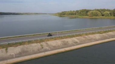 Bisikletçi, yaz günü nehir köprüsünden motosikletle geçiyor. Adam Lake Road 'da modern spor motosikletine hızlı biniyor. Motosikletli baraj yolunda bisikletiyle yarışıyor. Macera kavramı. Hava görüntüsü.