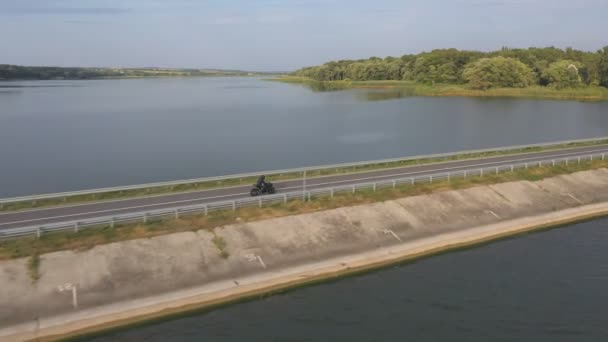 バイカーは夏の日に川の橋を通ってバイクを運転しています 湖の道路で現代のスポーツバイクに乗って高速男 オートバイ愛好家はダムのルートで自転車をレース 冒険の概念 空中射撃 — ストック動画