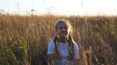 Beautiful smiling girl looking into camera against background of uncultivated meadow. Portrait of happy small kid straightens her blonde hair sitting on grass at field. Concept of resting. Dolly shot.