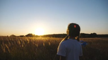 Sevimli bayan çocuk çim tarlasında yürürken oyuncak uçakla oynuyor. Kaygısız küçük çocuk gün batımında uçakla çayırların arasına gidiyor. Küçük kız özgürlüğün tadını çıkarıyor. Çocukluk hayali konsepti. Dolly vurdu..