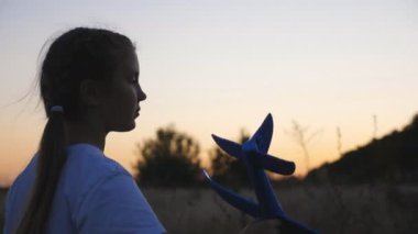 Alacakaranlıkta hareket eden oyuncak uçağıyla küçük şirin çocuk tutucularını yakın plan. Küçük kız kırsalda yürürken uçakla oynuyor. Tasasız çocuk yaz doğasında gezintiden zevk alıyor. Çocukluk hayali konsepti.