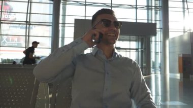 Havaalanında gülümseyen genç bir işadamı telefonda konuşuyor ve arka planda güneş parlaması var. Ofiste cep telefonuyla konuşan güneş gözlüklü mutlu iş adamının portresi. Yavaş çekim.