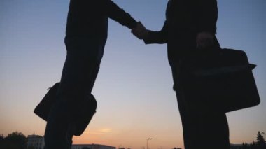 Evrak çantalı başarılı iş adamlarının şehir ortamında buluşup birbirlerini selamladıkları Dolly 'nin fotoğrafı. Gün batımında şehirde tokalaşan iki meslektaş. Dışarıda iş tokalaşması. Düşük açı görünümü.
