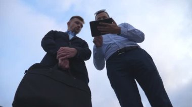 Genç iş adamı, şehirdeki meslektaşına tablet ekranında bir sunum gösteriyor. Başarılı girişimciler arka planda mavi gökyüzü olan dijital tablet üzerinde çalışıyorlar. Düşük açı görünümü Yavaş çekim.