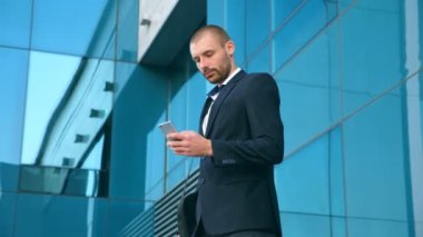 Ofisin yakınında akıllı telefon kullanan yakışıklı bir iş adamı. Genç iş adamı modern binanın yakınındaki akıllı telefondan haber okuyor. Dışarıda çalışan adamın portresi. Yavaş çekim. Yakın çekim..