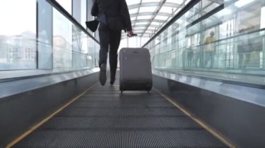 Terminal koridorundaki yürüyen merdivende yürüyen işadamlarının bacakları. Bavullu ve rulolu bir adamın arka görüntüsü uçuşa geç kalıyor. Takım elbiseli başarılı bir adam içeride koşuyor. İş gezisi için acele eden adam. Yavaş çekim.