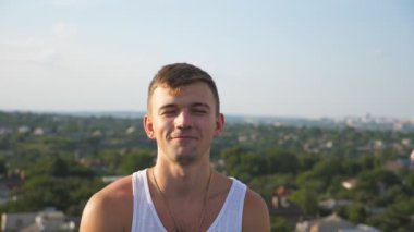 Portrait of young happy man sitting on rooftop at cityscape background. Handsome smiling guy looking into camera with joy expression on face. Sincere emotions of happy person. Dolly shot Slow motion.